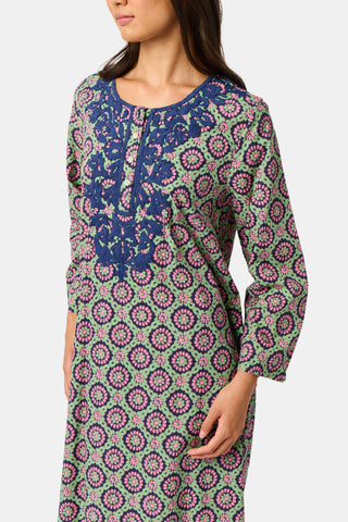 Wearing the Amelia Inez Long Kurta by Roller Rabbit, a woman with brown boots sits on the railing of a wooden porch, smiling, with a rustic building in the background.