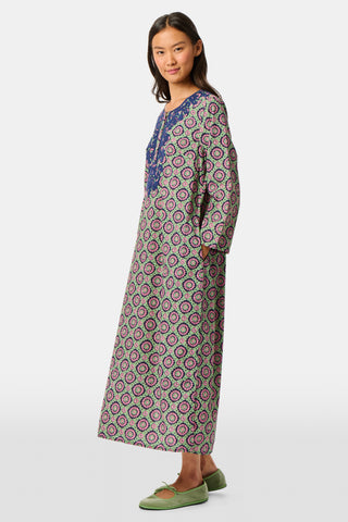 Wearing the Amelia Inez Long Kurta by Roller Rabbit, a woman with brown boots sits on the railing of a wooden porch, smiling, with a rustic building in the background.