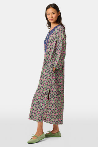 Wearing the Amelia Inez Long Kurta by Roller Rabbit, a woman with brown boots sits on the railing of a wooden porch, smiling, with a rustic building in the background.