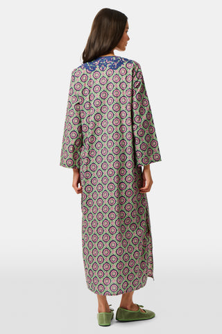Wearing the Amelia Inez Long Kurta by Roller Rabbit, a woman with brown boots sits on the railing of a wooden porch, smiling, with a rustic building in the background.