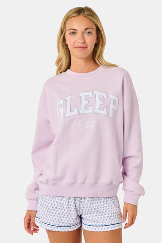 A woman wearing the Roller Rabbit Sleep Club Andie Sweatshirt in pastel lavender, paired with patterned white shorts, stands and smiles against a plain white background.