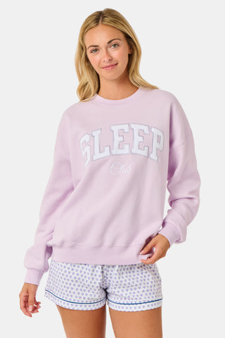 A woman wearing the Roller Rabbit Sleep Club Andie Sweatshirt in pastel lavender, paired with patterned white shorts, stands and smiles against a plain white background.
