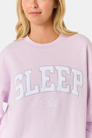 A woman wearing the Roller Rabbit Sleep Club Andie Sweatshirt in pastel lavender, paired with patterned white shorts, stands and smiles against a plain white background.
