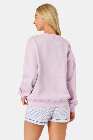 A woman wearing the Roller Rabbit Sleep Club Andie Sweatshirt in pastel lavender, paired with patterned white shorts, stands and smiles against a plain white background.