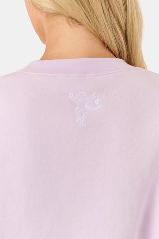 A woman wearing the Roller Rabbit Sleep Club Andie Sweatshirt in pastel lavender, paired with patterned white shorts, stands and smiles against a plain white background.