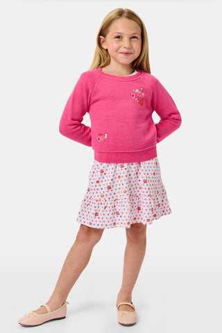 A young girl smiles, wearing the Roller Rabbit Kids Autumn Hearts Crewneck—a pink sweatshirt featuring small embroidered butterflies—paired with a white skirt adorned with a colorful heart pattern, as she stands with her hands behind her back.