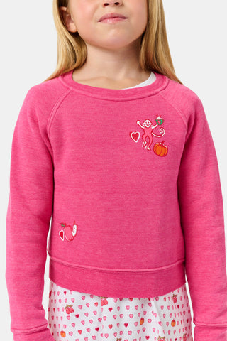 A young girl smiles, wearing the Roller Rabbit Kids Autumn Hearts Crewneck—a pink sweatshirt featuring small embroidered butterflies—paired with a white skirt adorned with a colorful heart pattern, as she stands with her hands behind her back.