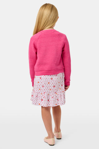 A young girl smiles, wearing the Roller Rabbit Kids Autumn Hearts Crewneck—a pink sweatshirt featuring small embroidered butterflies—paired with a white skirt adorned with a colorful heart pattern, as she stands with her hands behind her back.