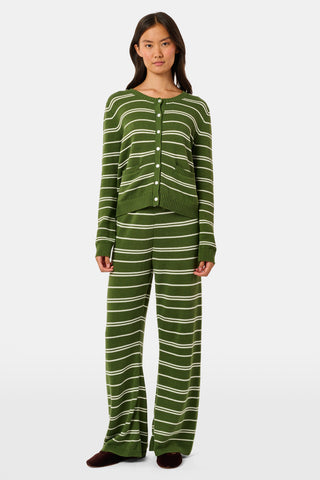 A person wearing the Roller Rabbit Autumn Stripe Olivia Pant—green and white striped, pull-on, flared—and a matching sweater lifts the hem to reveal their midriff. Brown slippers are visible. The background is white.