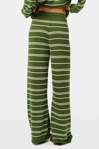 A person wearing the Roller Rabbit Autumn Stripe Olivia Pant—green and white striped, pull-on, flared—and a matching sweater lifts the hem to reveal their midriff. Brown slippers are visible. The background is white.