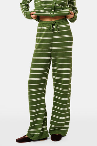 A person wearing the Roller Rabbit Autumn Stripe Olivia Pant—green and white striped, pull-on, flared—and a matching sweater lifts the hem to reveal their midriff. Brown slippers are visible. The background is white.
