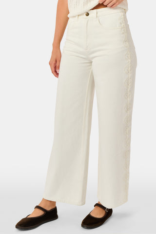 Wearing Roller Rabbit’s Elsa Eyelet Danielle Jean—a high-waisted, wide-leg style in ecru with delicate scalloped lace trim along the sides—paired with a white top and black Mary Jane shoes, the individual stands against a plain white background.