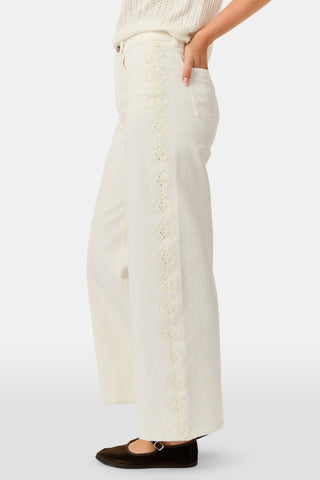 Wearing Roller Rabbit’s Elsa Eyelet Danielle Jean—a high-waisted, wide-leg style in ecru with delicate scalloped lace trim along the sides—paired with a white top and black Mary Jane shoes, the individual stands against a plain white background.