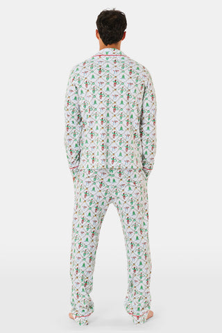 A woman wears a SERFLEX Deck The Halls Bailey Pajamas set featuring a short-sleeved top and shorts with a white background and a repeating Christmas-themed pattern of elves, snowmen, and evergreen trees.