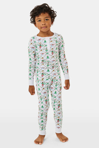 A child wearing SERFLEX Kids Deck The Halls Pajamas, featuring a repeating holiday pattern of snowmen, Christmas trees, and elves connected by garland on long-sleeved Pima cotton.