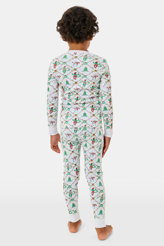 A child wearing SERFLEX Kids Deck The Halls Pajamas, featuring a repeating holiday pattern of snowmen, Christmas trees, and elves connected by garland on long-sleeved Pima cotton.