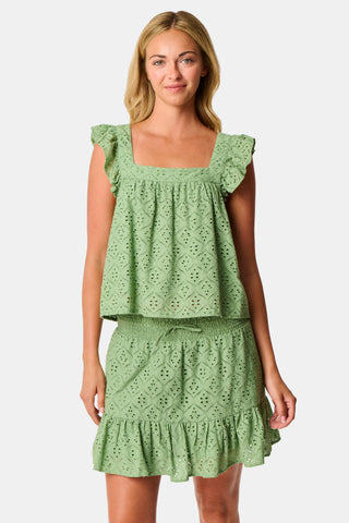 A person wearing the Roller Rabbit Elsa Eyelet Peyton Skirt in sage green, featuring a cotton eyelet fabric, ruffled hem, and elastic waistband, paired with a matching top and slightly lifting the top to reveal the skirt’s waist.