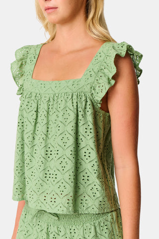 A person wearing the Roller Rabbit Elsa Eyelet Peyton Skirt in sage green, featuring a cotton eyelet fabric, ruffled hem, and elastic waistband, paired with a matching top and slightly lifting the top to reveal the skirt’s waist.