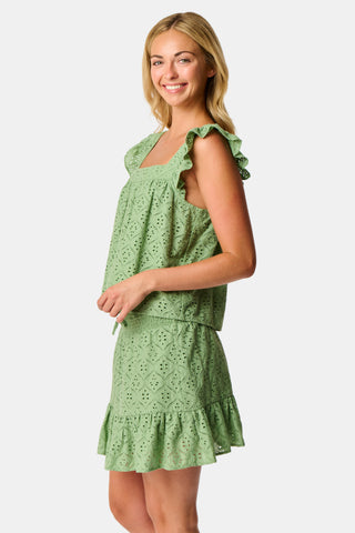 A person wearing the Roller Rabbit Elsa Eyelet Peyton Skirt in sage green, featuring a cotton eyelet fabric, ruffled hem, and elastic waistband, paired with a matching top and slightly lifting the top to reveal the skirt’s waist.