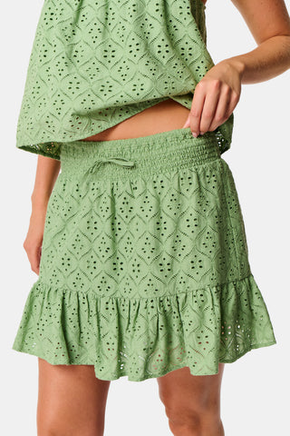 A person wearing the Roller Rabbit Elsa Eyelet Peyton Skirt in sage green, featuring a cotton eyelet fabric, ruffled hem, and elastic waistband, paired with a matching top and slightly lifting the top to reveal the skirt’s waist.