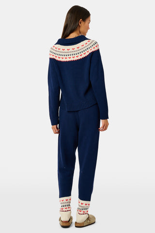 A person leans against a wooden fence wearing a matching navy blue Fairisle sweater, scarf, and INKASIGN Fairisle Hearts Jett Pant, all featuring heart and geometric patterns for an apres-ski glam look.