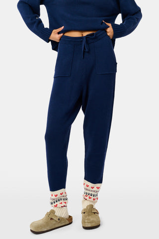 A person leans against a wooden fence wearing a matching navy blue Fairisle sweater, scarf, and INKASIGN Fairisle Hearts Jett Pant, all featuring heart and geometric patterns for an apres-ski glam look.