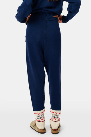 A person leans against a wooden fence wearing a matching navy blue Fairisle sweater, scarf, and INKASIGN Fairisle Hearts Jett Pant, all featuring heart and geometric patterns for an apres-ski glam look.