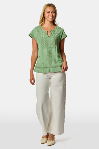 A woman wearing the Roller Rabbit Faith Top—a light green, short-sleeve embroidered cotton blouse with a split neckline—paired with white pants, stands against a plain light background.