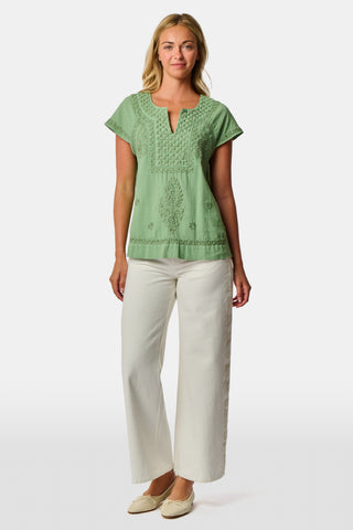 A woman wearing the Roller Rabbit Faith Top—a light green, short-sleeve embroidered cotton blouse with a split neckline—paired with white pants, stands against a plain light background.