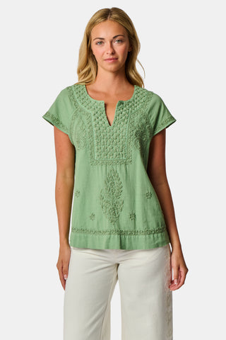 A woman wearing the Roller Rabbit Faith Top—a light green, short-sleeve embroidered cotton blouse with a split neckline—paired with white pants, stands against a plain light background.