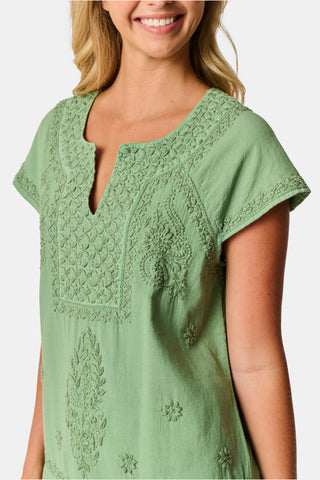 A woman wearing the Roller Rabbit Faith Top—a light green, short-sleeve embroidered cotton blouse with a split neckline—paired with white pants, stands against a plain light background.