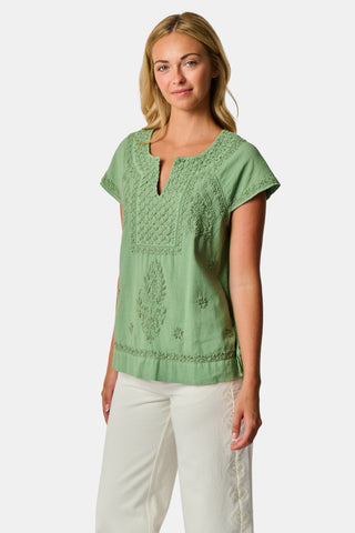 A woman wearing the Roller Rabbit Faith Top—a light green, short-sleeve embroidered cotton blouse with a split neckline—paired with white pants, stands against a plain light background.