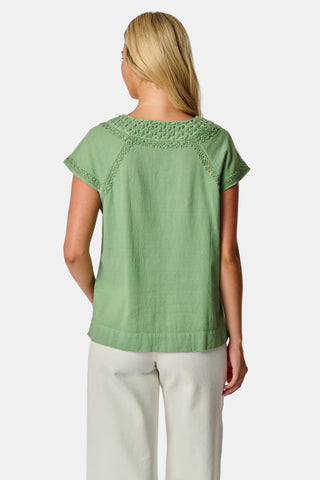 A woman wearing the Roller Rabbit Faith Top—a light green, short-sleeve embroidered cotton blouse with a split neckline—paired with white pants, stands against a plain light background.