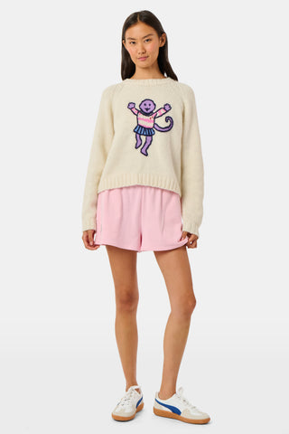 A young woman sits on a bench, wearing the Roller Rabbit Game Day Brittany Sweater with a cartoon figure, layered over a light dress. She is beside a blue patterned backpack and surrounded by greenery.