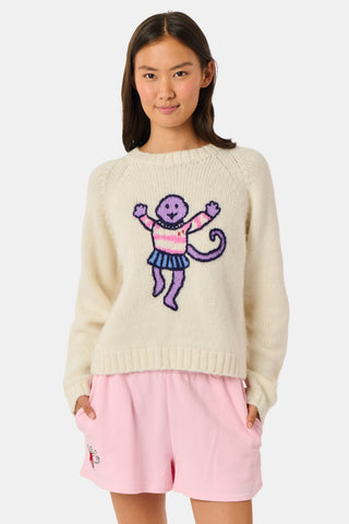A young woman sits on a bench, wearing the Roller Rabbit Game Day Brittany Sweater with a cartoon figure, layered over a light dress. She is beside a blue patterned backpack and surrounded by greenery.
