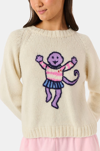 A young woman sits on a bench, wearing the Roller Rabbit Game Day Brittany Sweater with a cartoon figure, layered over a light dress. She is beside a blue patterned backpack and surrounded by greenery.