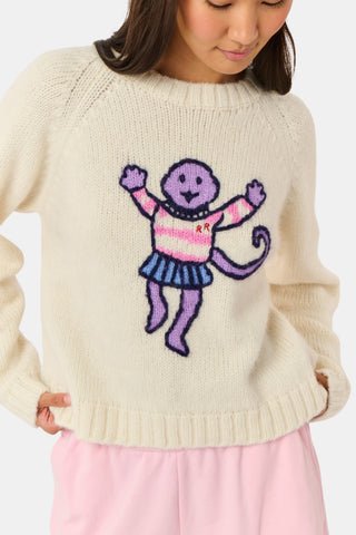 A young woman sits on a bench, wearing the Roller Rabbit Game Day Brittany Sweater with a cartoon figure, layered over a light dress. She is beside a blue patterned backpack and surrounded by greenery.