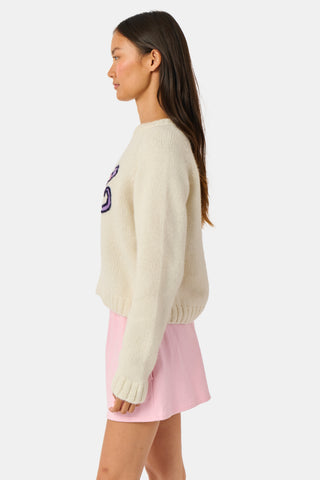 A young woman sits on a bench, wearing the Roller Rabbit Game Day Brittany Sweater with a cartoon figure, layered over a light dress. She is beside a blue patterned backpack and surrounded by greenery.