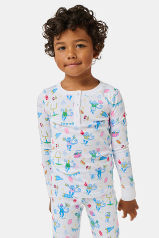 The Kids Game Day Pajamas by Roller Rabbit are a children’s long-sleeve pajama set crafted from soft Pima cotton, featuring a vibrant party-themed print with colorful dinosaurs, bunting, gifts, and party hats on a white background.