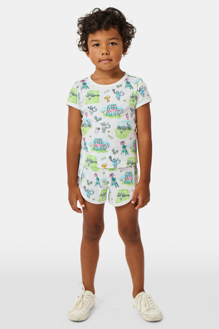 A girl stands outdoors in a Roller Rabbit Kids Game, Set, Monkey Playa Play Set, holding out a tennis ball toward the camera. Her playful outfit is perfect for kids’ tennis activities, and the photo is taken from a low angle with blue sky and trees in the background.