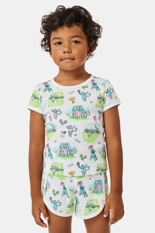 A girl stands outdoors in a Roller Rabbit Kids Game, Set, Monkey Playa Play Set, holding out a tennis ball toward the camera. Her playful outfit is perfect for kids’ tennis activities, and the photo is taken from a low angle with blue sky and trees in the background.