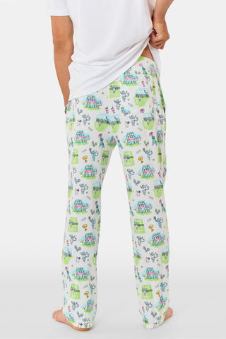 The Roller Rabbit Mens Game, Set, Monkey Troy Pajamas feature a white short-sleeve t-shirt paired with white pajama pants decorated with a vibrant dinosaur and jungle print in green, blue, and black. This playful Pima cotton loungewear set from Roller Rabbit is displayed on a plain white background.