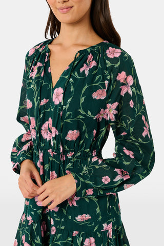 A model wears the SOPHISTICA Greta Floral Belle Dress, a dark green cotton mini dress featuring a pink floral print, a gathered waist, and a ruffled hem.