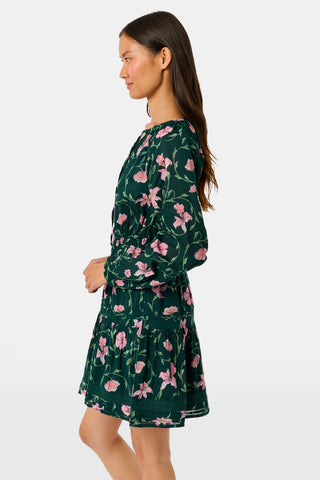 A model wears the SOPHISTICA Greta Floral Belle Dress, a dark green cotton mini dress featuring a pink floral print, a gathered waist, and a ruffled hem.