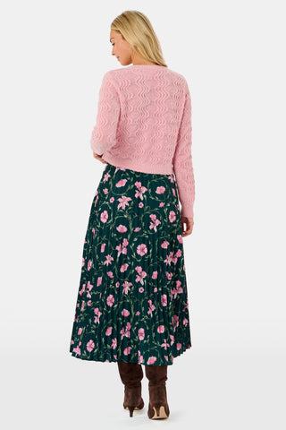 The SOPHISTICA Greta Floral Megan Skirt is a midi-length pleated skirt featuring a dark green background with a pink floral pattern, worn with a pink cable-knit sweater and brown suede ankle boots.