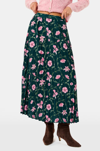 The SOPHISTICA Greta Floral Megan Skirt is a midi-length pleated skirt featuring a dark green background with a pink floral pattern, worn with a pink cable-knit sweater and brown suede ankle boots.