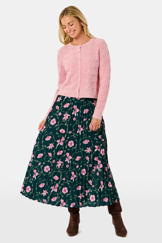 The SOPHISTICA Greta Floral Megan Skirt is a midi-length pleated skirt featuring a dark green background with a pink floral pattern, worn with a pink cable-knit sweater and brown suede ankle boots.