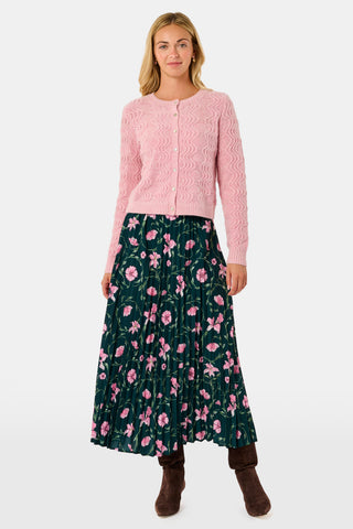 The SOPHISTICA Greta Floral Megan Skirt is a midi-length pleated skirt featuring a dark green background with a pink floral pattern, worn with a pink cable-knit sweater and brown suede ankle boots.