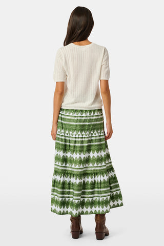 A person wearing the Roller Rabbit Hayam Carlisa Skirt in an olive and white print, paired with a cream knit top and brown boots, stands against a plain white background.