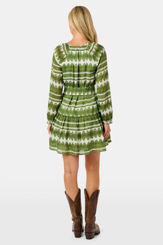 A woman outdoors wears the Hayam Monroe Dress by Roller Rabbit—an olive and white long-sleeved dress with a matching belt. She walks with another person, with trees and a wooden fence in the background.